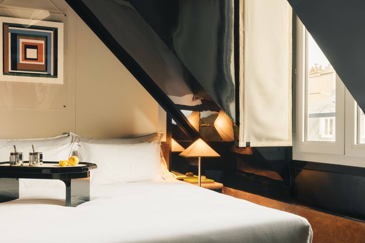 A modern bedroom with a large bed, a bedside table with a lamp, and a view of the city through a window.