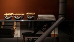 A modern office chair with a wooden base and backrest, positioned in front of a reflective black desk with a stack of white towels and a black cylindrical object.
