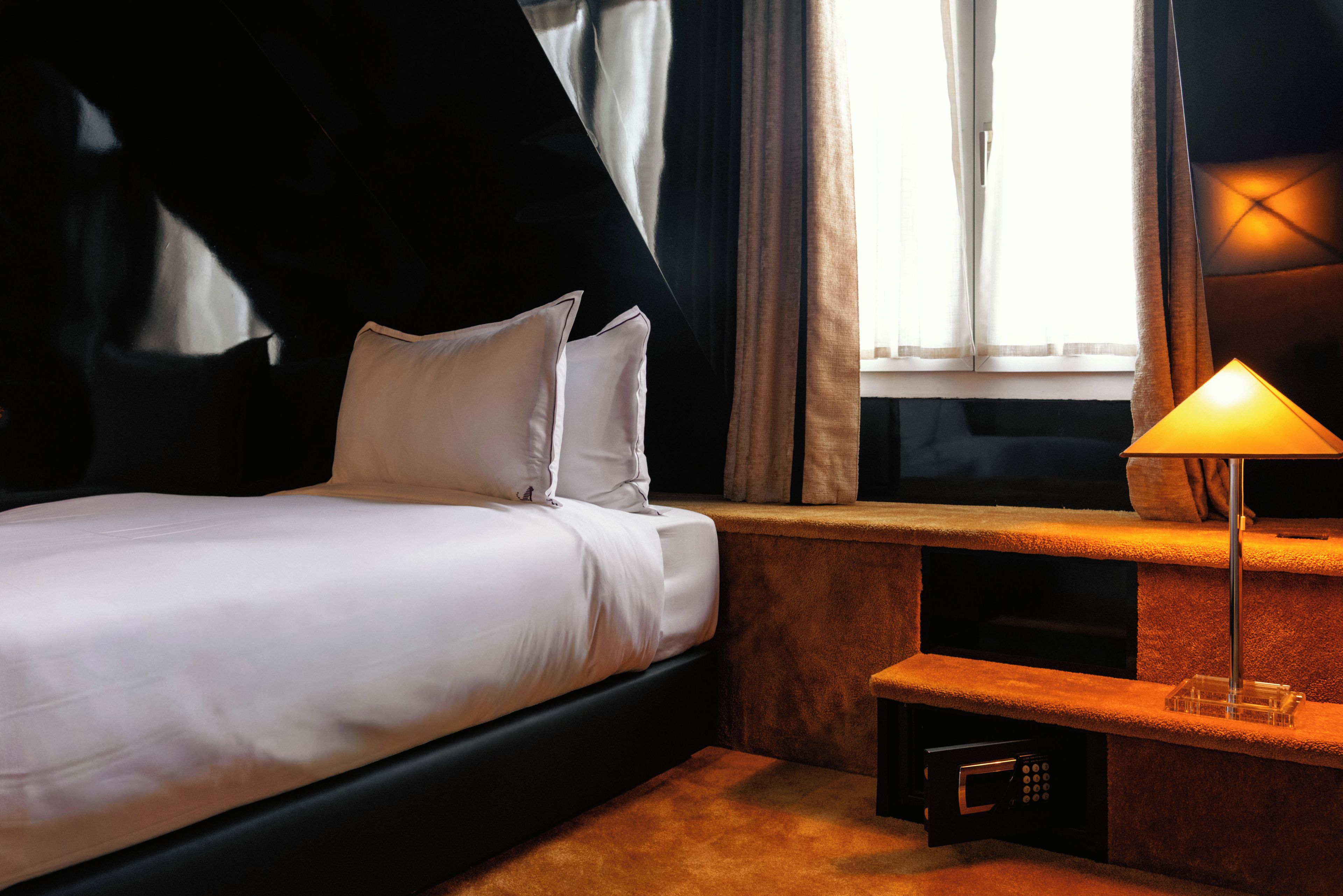 A hotel room with a bed, a bedside table with a lamp, a window with curtains, and a wall-mounted TV.