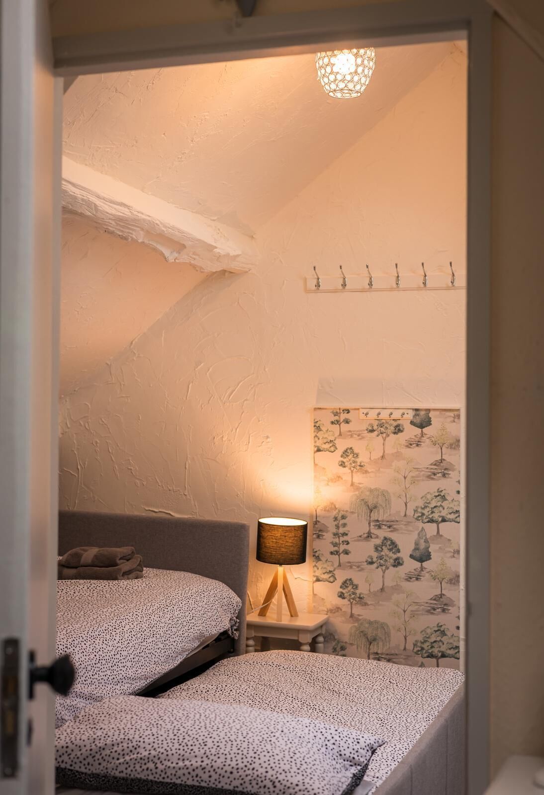 Un dormitorio con techo inclinado, una cama con edredón estampado, una lámpara de noche y un estante con ganchos.