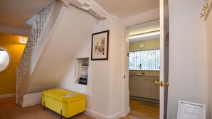 Une pièce avec un pouf jaune, un tableau encadré au mur, un escalier et une porte donnant sur une cuisine avec une fenêtre.