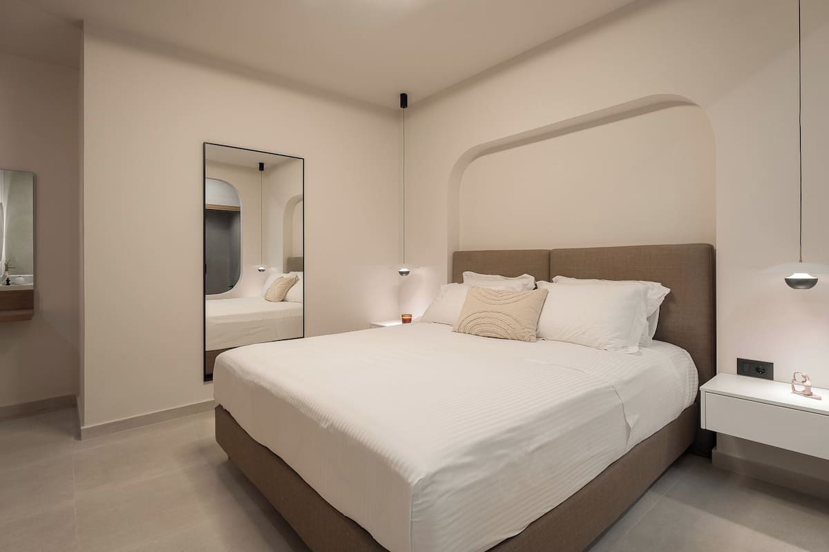A modern bedroom with a large bed, a bedside table, a nightstand, a mirror, and a wall-mounted light fixture.