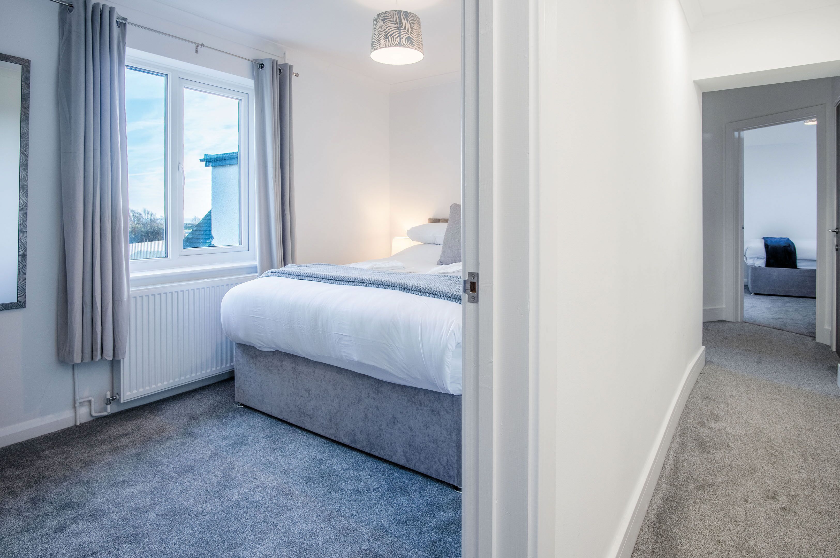 A bedroom with a bed, a window with curtains, a radiator, and a hallway leading to another room.