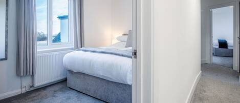A bedroom with a bed, a window with curtains, a radiator, and a hallway leading to another room.