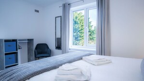 Ein ordentlich bezogenes Bett mit weißen Leinen und einer grauen Decke, ein blauer Kleiderschrank, ein Stuhl und ein Fenster mit Vorhängen.