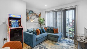 A living room with a blue sectional sofa, a retro arcade game, a large abstract painting, and a view of the city through floor-to-ceiling windows. - 2 Bed Apt Sleeps 7 - Arcade Machine, Wifi, Central (Birmingham)