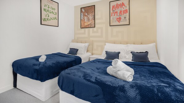 Quarto de hotel com duas camas, cada uma com um cobertor azul e almofadas brancas. Há quadros na parede, um com o texto "It's not Versacity, it's Versace" e outro com "Mama I'm Rich".