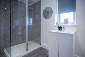 Un baño moderno con ducha envuelta en vidrio, un lavabo blanco con espejo y una ventana con persiana azul.