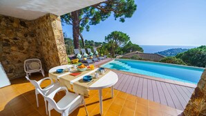Un área de comedor al aire libre iluminada por el sol, con piscina y vista al océano.