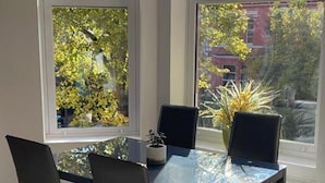 A dining area with a table and four chairs, large windows with a view of trees and buildings, and a radiator on the floor. - Spacious 2-bed Queens by CityGo (Belfast)