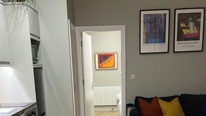 A modern kitchen with white cabinets and a built-in oven, a dining area with a glass table and chairs, and a living room with a dark sofa and colorful cushions. - CityGo Apartments Queens (Belfast)