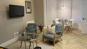 A living room with two armchairs, a glass coffee table, a TV mounted on the wall, a dining table with chairs, and a potted plant. - CityGo Apartments Queens (Belfast)