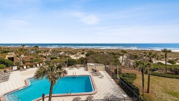 A swimming pool with a view of the ocean, surrounded by palm trees and a fenced area with lounge chairs. - Oceanfront 2BR condo with pool, tennis, balcony, and beach access. (Fernandina Beach)