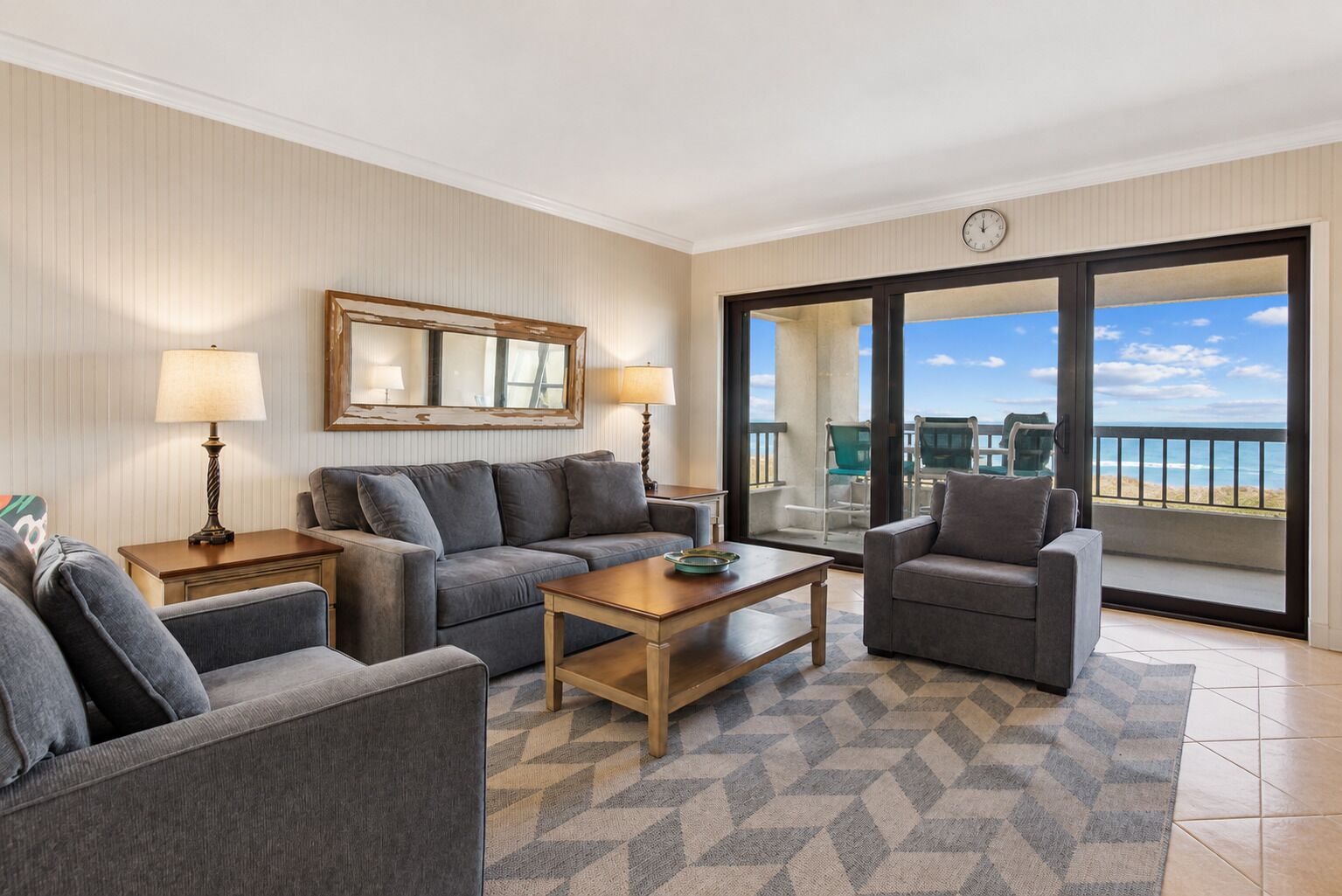 A modern living room with a sofa, armchair, and coffee table. There is a large window with a view of the ocean.