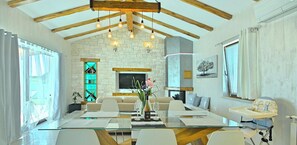 Un salon moderne avec une table à manger en verre, des canapés blancs, un téléviseur et un mur en pierre.