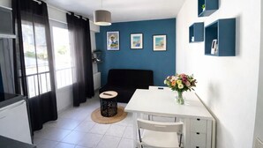 A modern room with a black sofa, a white dining table, and a small round table with a remote control. There are framed pictures on the wall and a vase with flowers on the dining table.