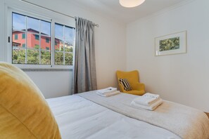 Una habitación de hotel con cama, ventana con cortinas, una silla amarilla y un cuadro en la pared.
