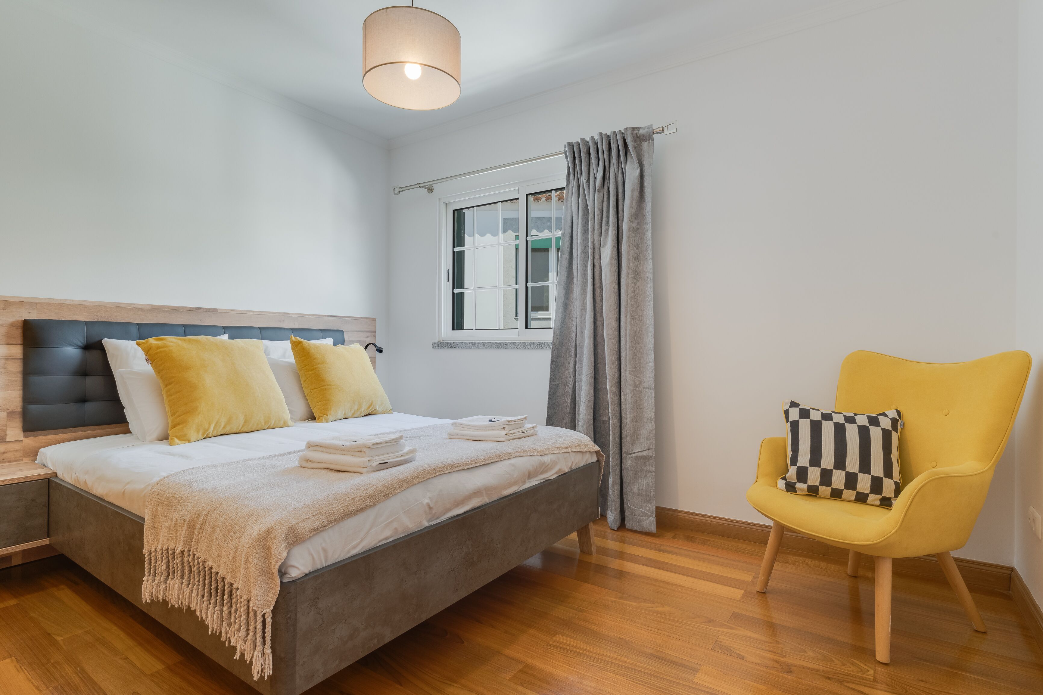 A bedroom with a bed, a chair, a window, and a hanging light fixture.