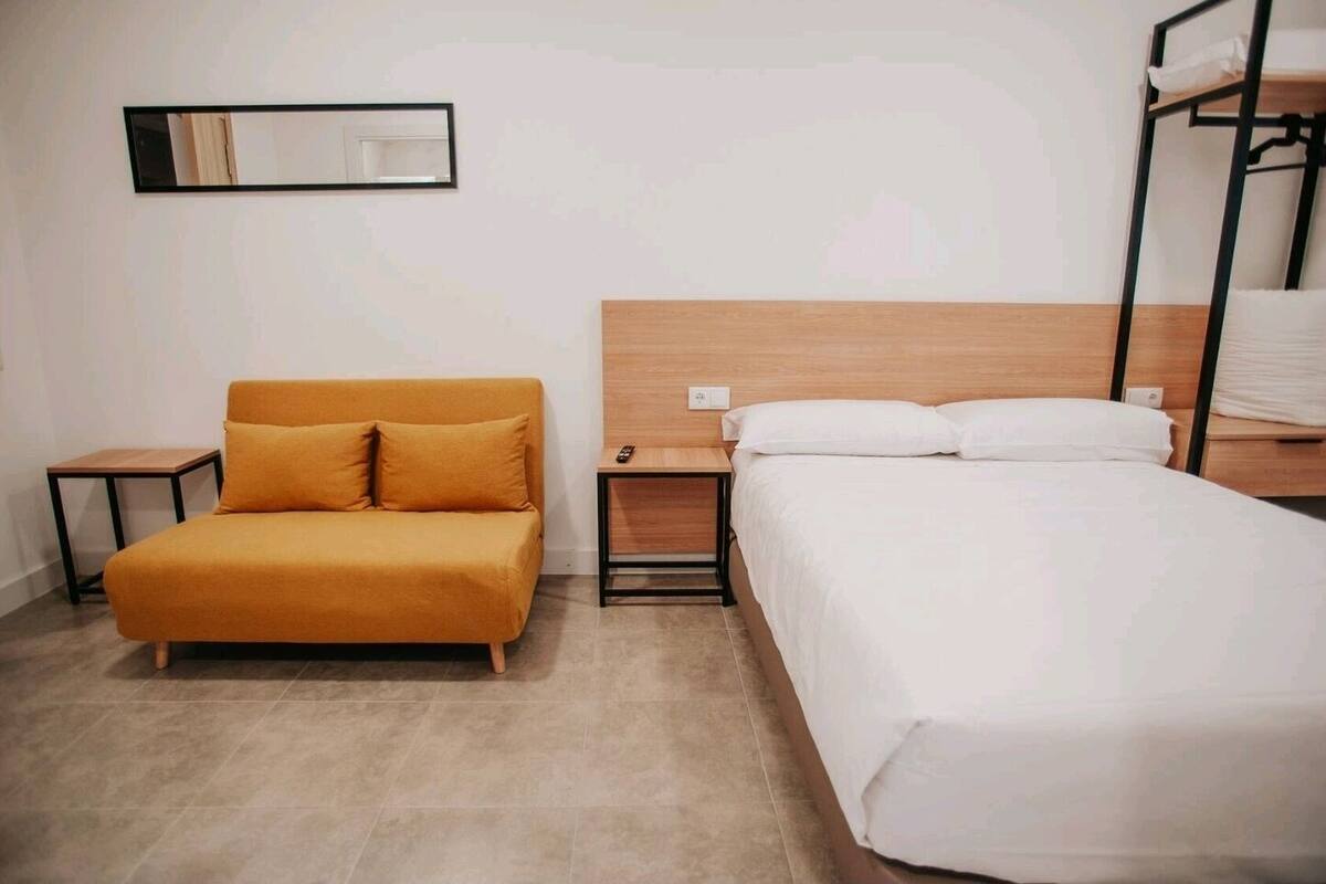 A hotel room with a sofa, a bed, a nightstand, a bedside table, a mirror, and a bunk bed.