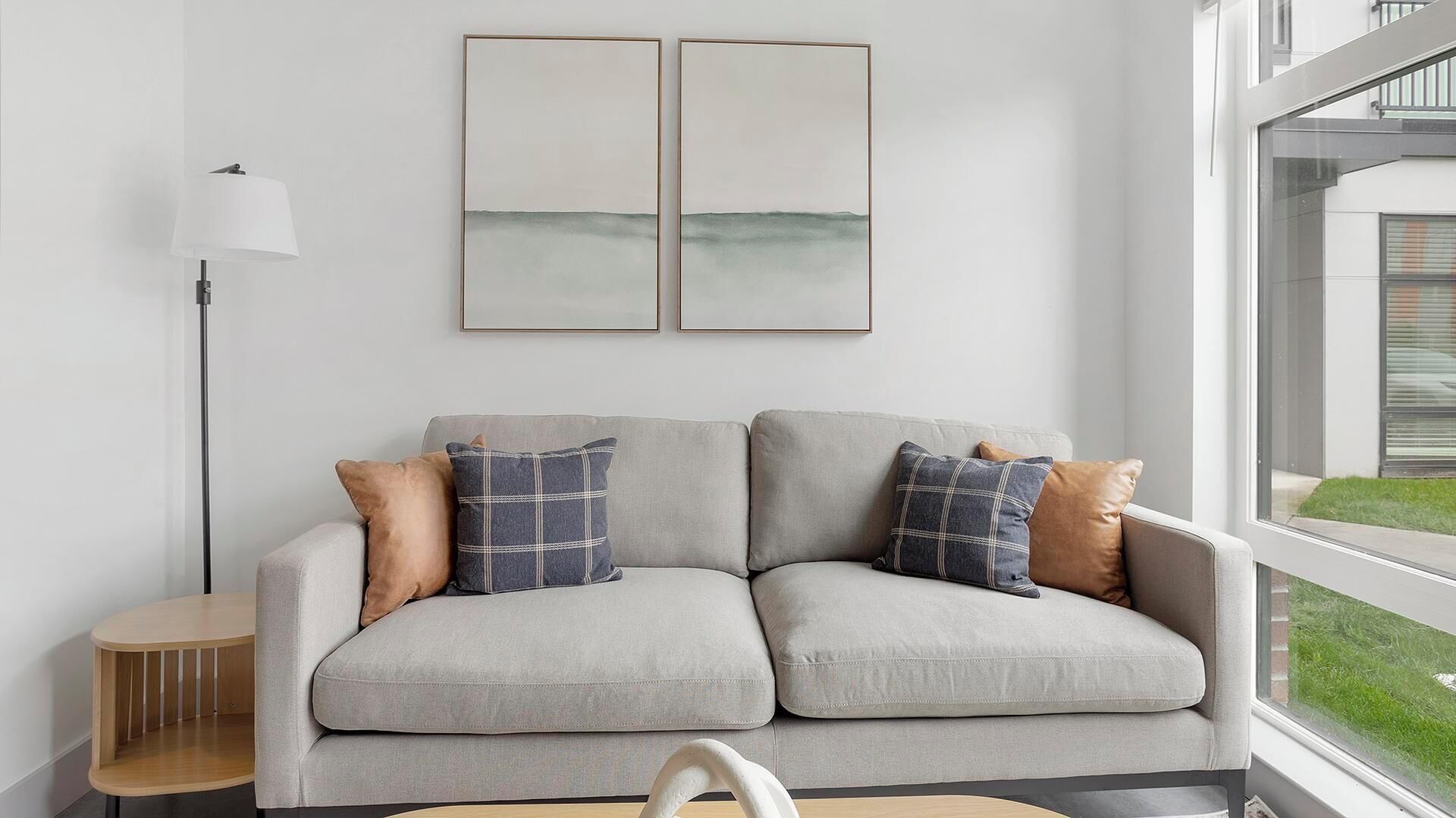 Een moderne woonkamer met een grijze bank, twee ingelijste kunstwerken erboven, een vloerlamp en uitzicht op een groen landschap door een raam.