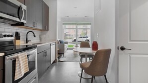 Eine moderne Küche mit Edelstahlgeräten, einem Essbereich mit rundem Tisch und zwei Stühlen sowie Blick auf die Straße durch große Fenster.