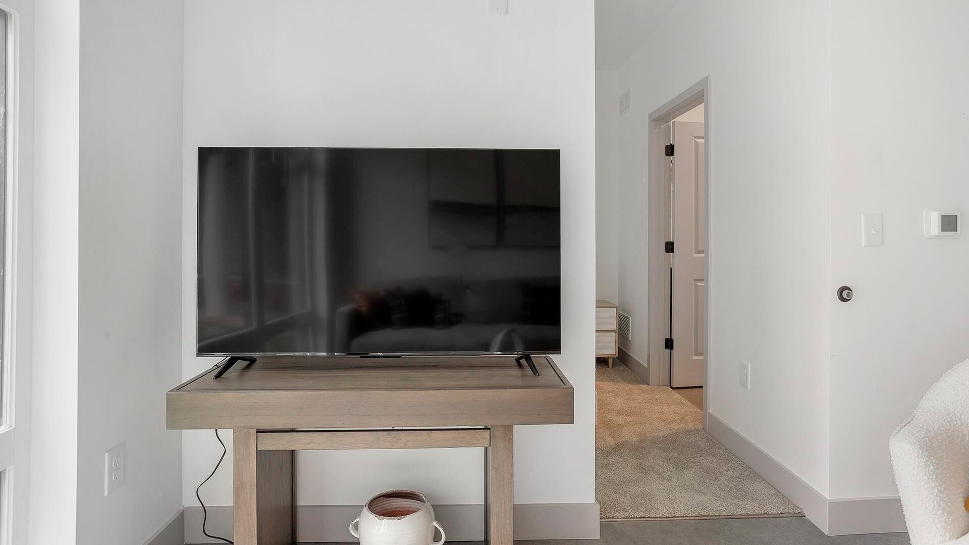 A modern living room with a large flat-screen TV on a wooden stand, a white mug on the floor, and a hallway leading to a bedroom.