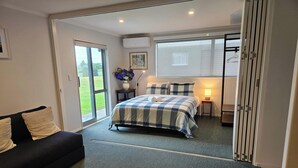 A bedroom with a bed, a sofa, a small table, a lamp, and a window. - Oceanview on Broadway (Waihi Beach)