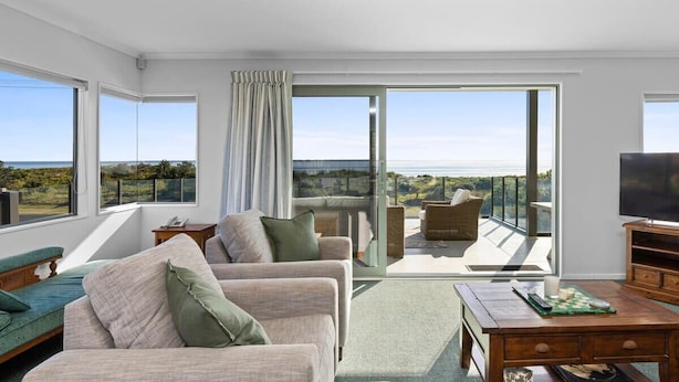 A modern living room with a sofa, a coffee table, and a TV. There is a balcony with a view of the landscape. - Oceanview on Broadway (Waihi Beach)