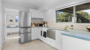 A modern kitchen with white cabinetry, stainless steel appliances, and a large window offering a view of trees. - Oceanview on Broadway (Waihi Beach)