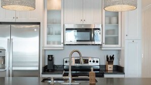A modern kitchen with white cabinetry, stainless steel appliances, a central island with a sink, and two wicker chairs. - Allure by Mira-Mar (Wrightsville Beach)