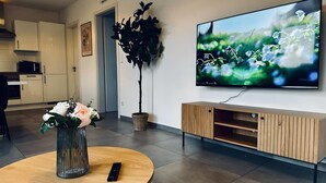 Ein modernes Wohnzimmer mit Couchtisch aus Holz, einem an der Wand montierten Fernseher, einer Topfpflanze und einem schlichten Schrank.