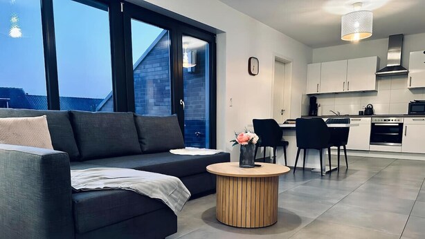 Ein modernes Wohnzimmer mit einem dunkelgrauen Sofa, einem runden Couchtisch aus Holz und einem Essbereich mit einem schwarzen Stuhl und weißen Schränken.