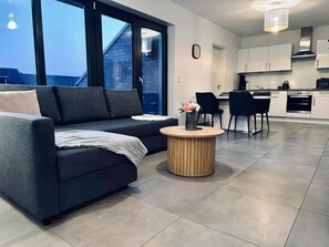 Een moderne woonkamer met een donkere bank, een ronde houten salontafel en een uitzicht op de lucht door grote ramen.