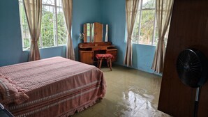 Ein Schlafzimmer mit einem Bett, einem Holztisch, einem Stuhl, einem Spiegel und einem Standventilator.