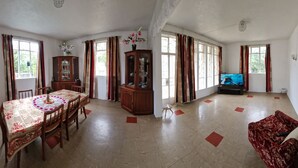 Ein geräumiges Wohnzimmer mit Essbereich, großen Fenstern mit Vorhängen, einem Fernseher und einem roten, gemusterten Sofa.
