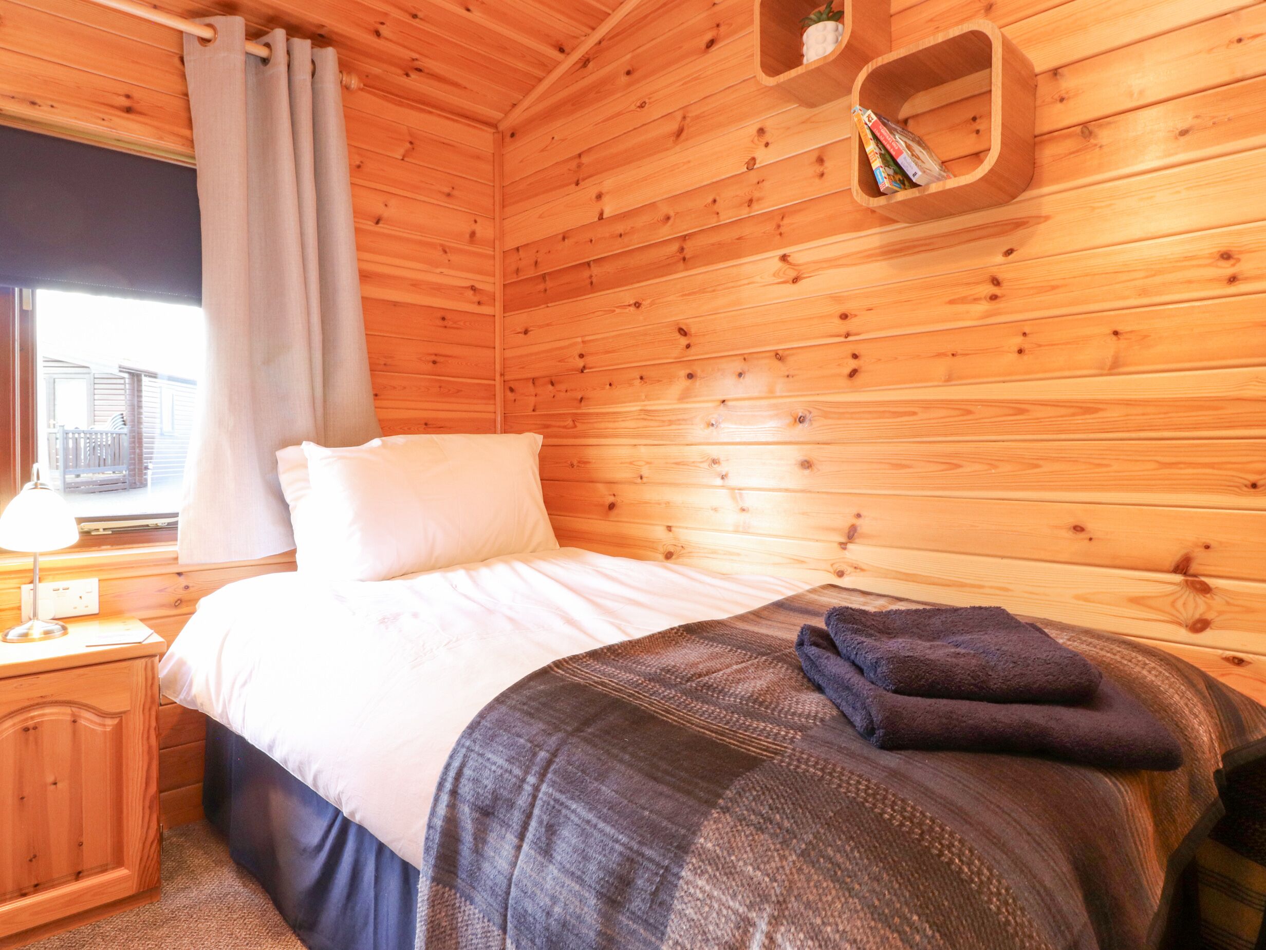 A wooden cabin room with a bed, a nightstand, a window with curtains, and a shelf with books.