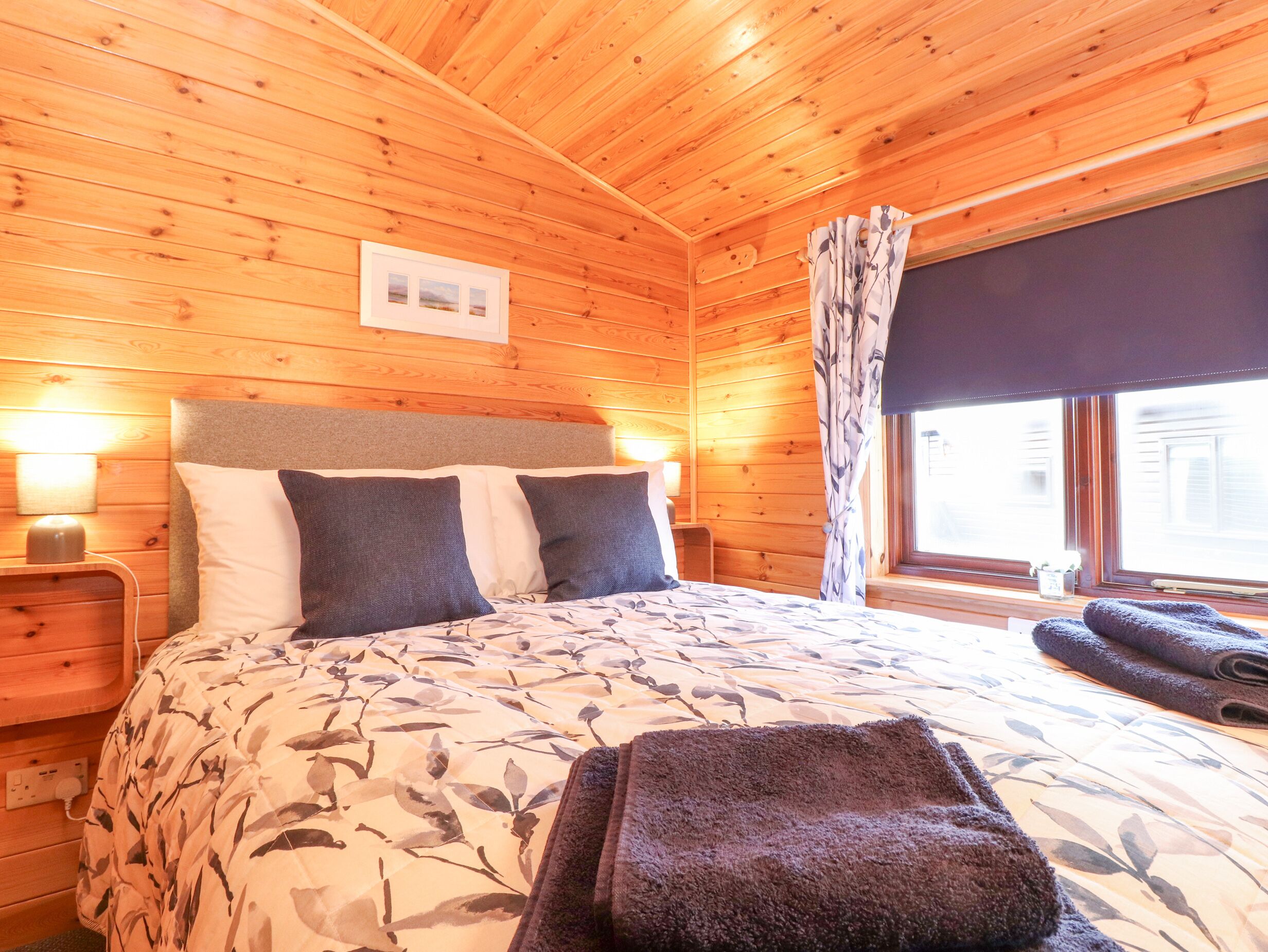 A wooden cabin bedroom with a bed, two bedside tables, a window with curtains, and a wall-mounted picture.