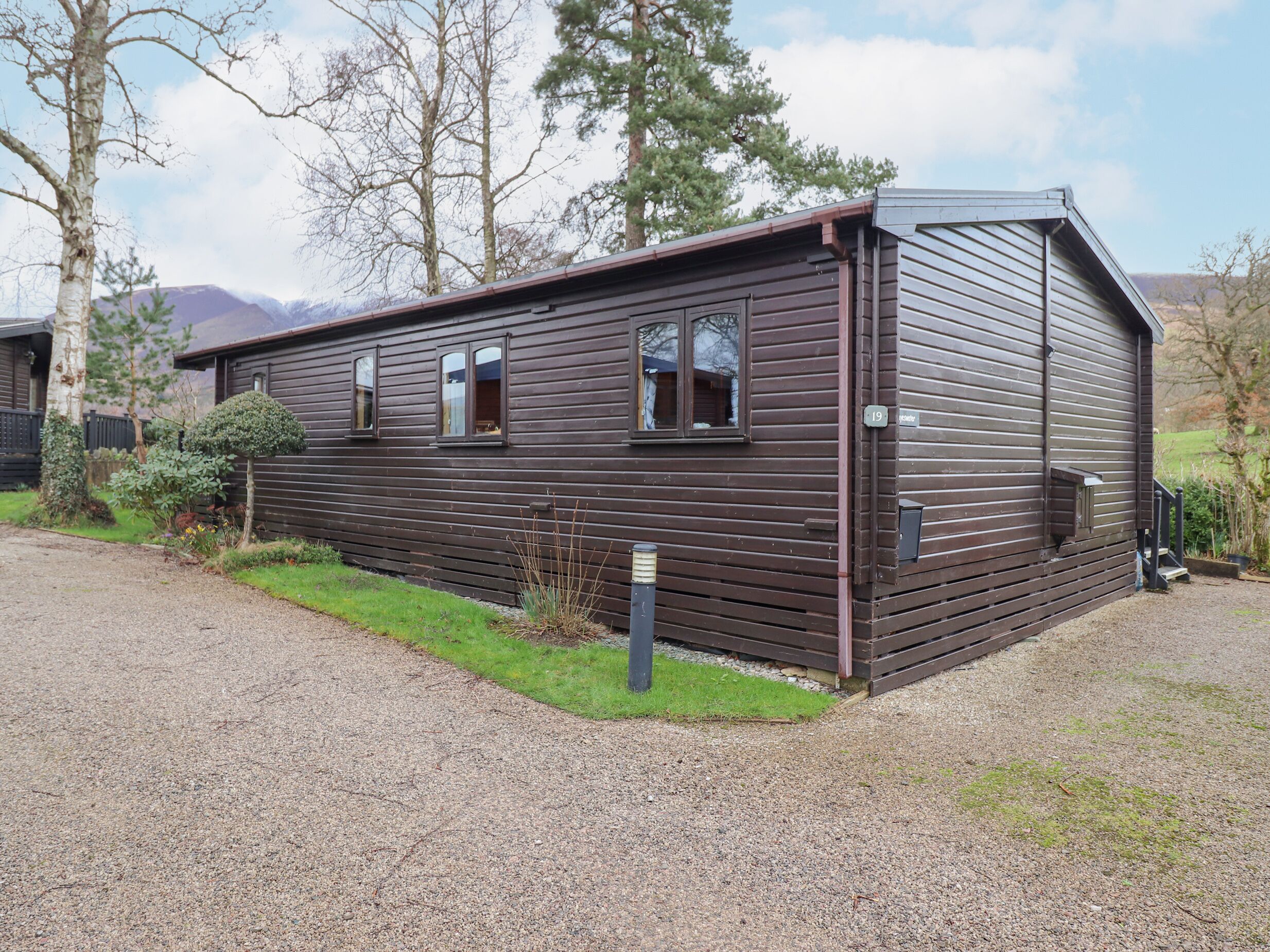 A single-story wooden cabin with a number 19 sign, a gravel driveway, and a small garden area.
