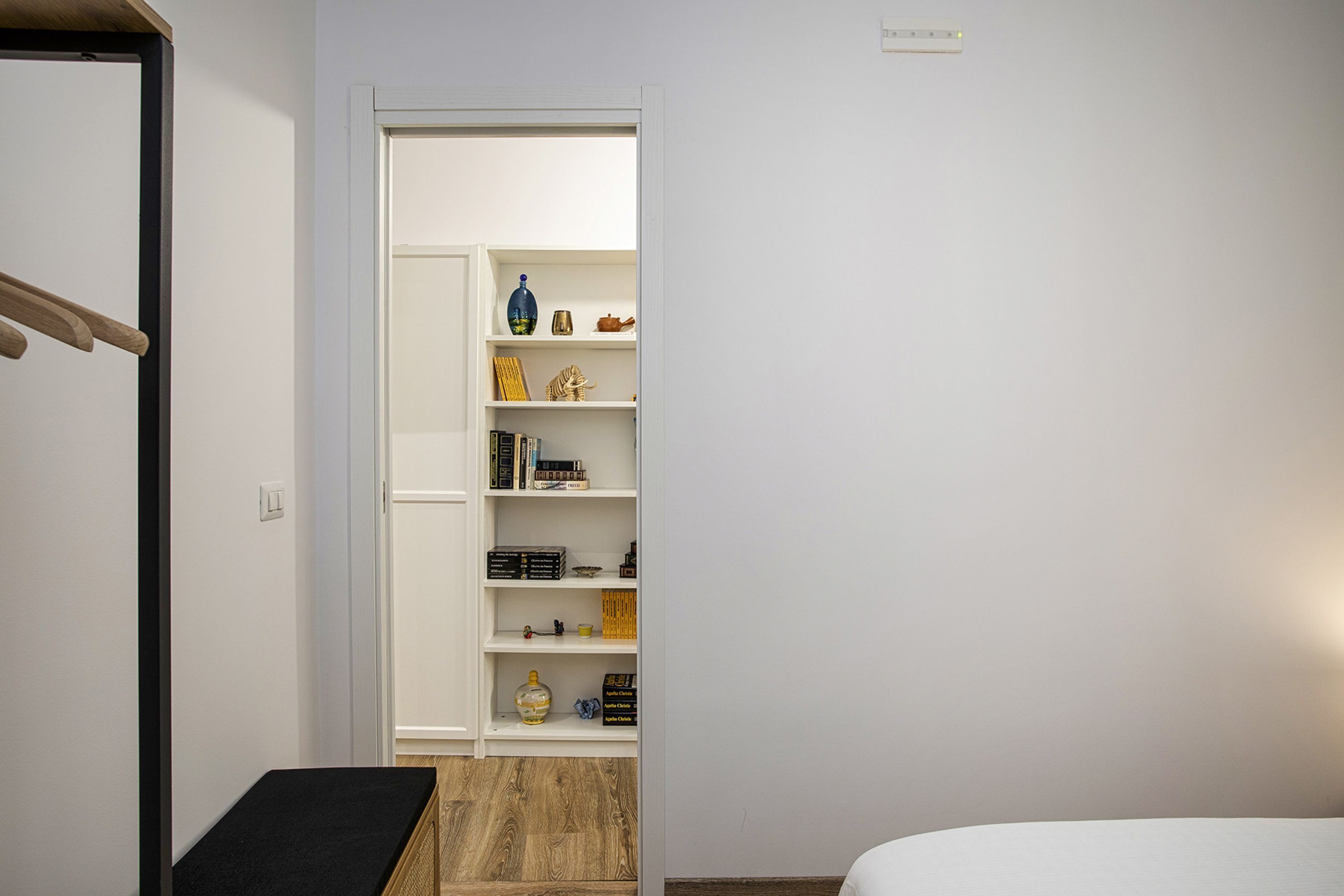 A bedroom with a bed, a wooden bench, a white wardrobe, and a shelf with various items.