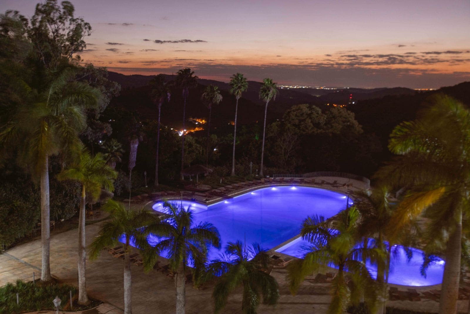 A nighttime view of a swimming pool surrounded by palm trees and a mountainous landscape.
