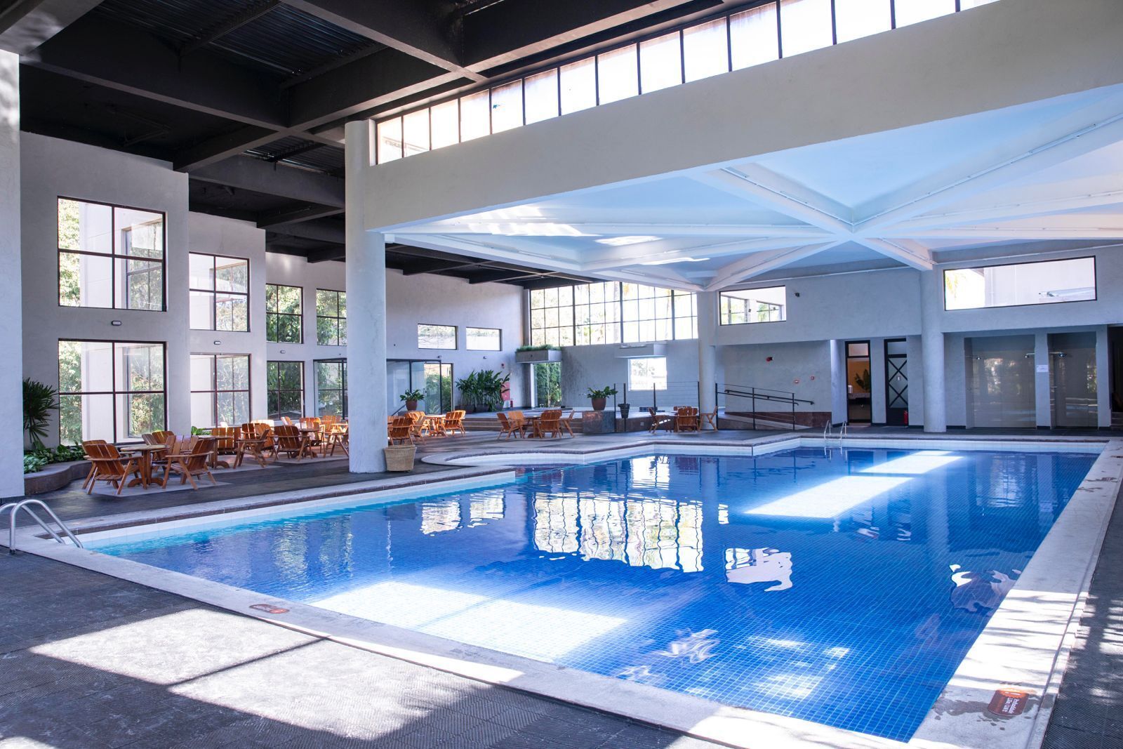 An indoor swimming pool with clear glass walls, multiple seating areas, and a high ceiling.