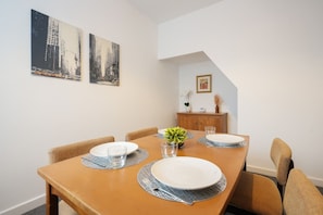 Un comedor con una mesa de madera dispuesta para cuatro personas, dos cuadros en la pared y una planta pequeña en la mesa.