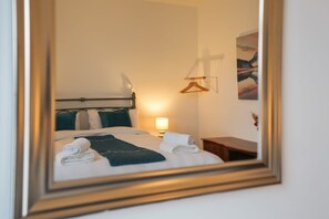 A hotel room with a bed, a bedside table, a lamp, a hanging coat rack, and a painting on the wall.
