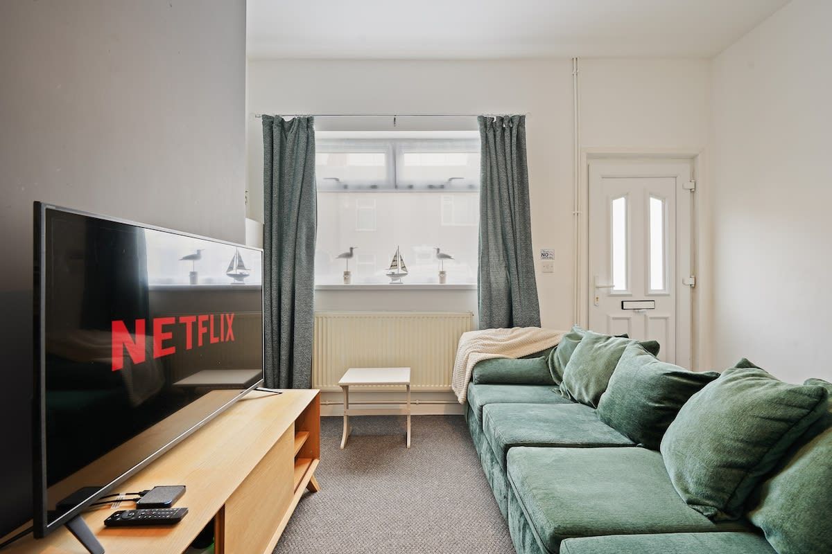 Un salon avec un téléviseur affichant le logo de Netflix, un canapé vert, une petite table en bois et une fenêtre avec des rideaux.
