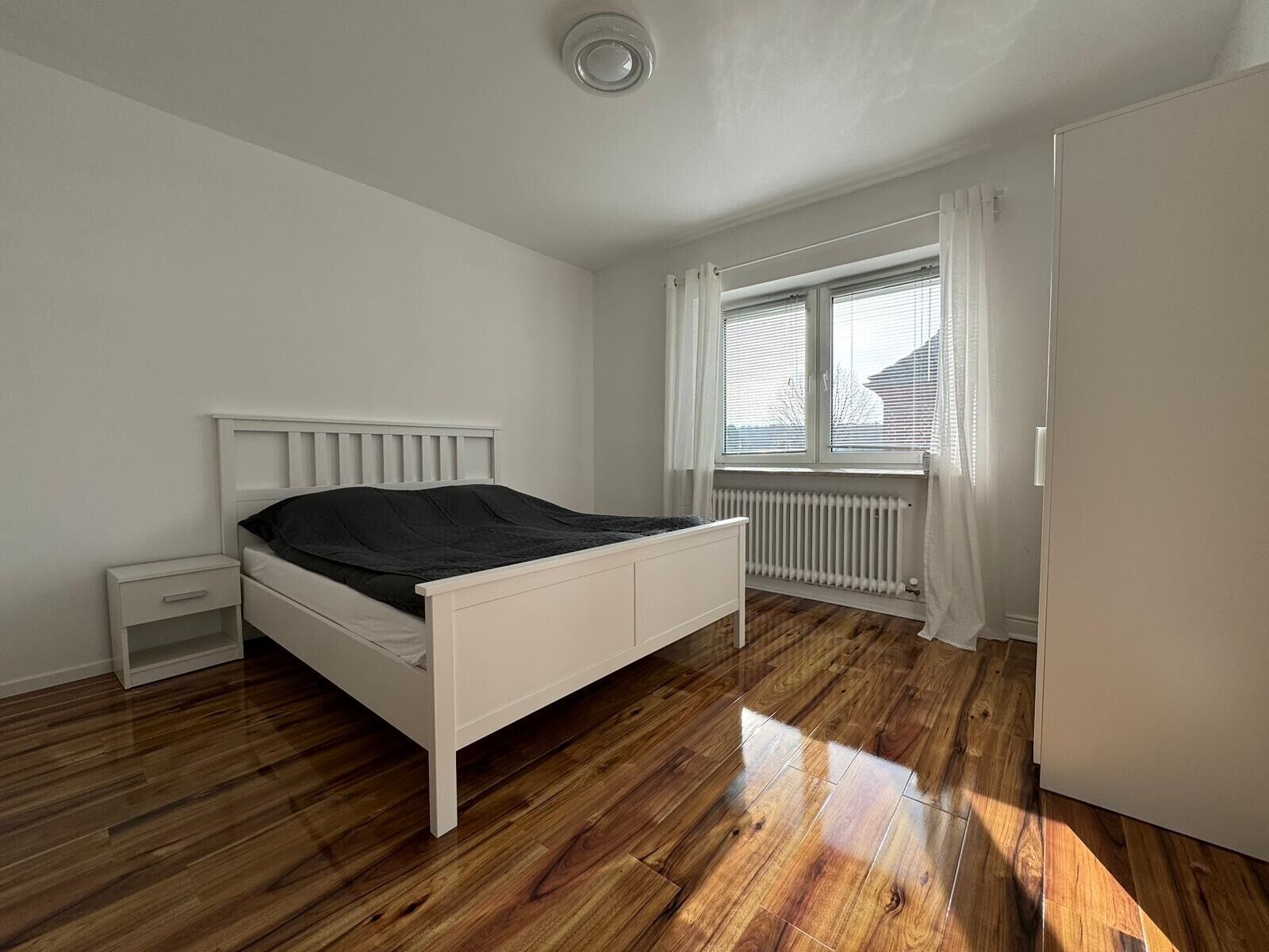 A bedroom with a white bed, a nightstand, a window with curtains, and a radiator.