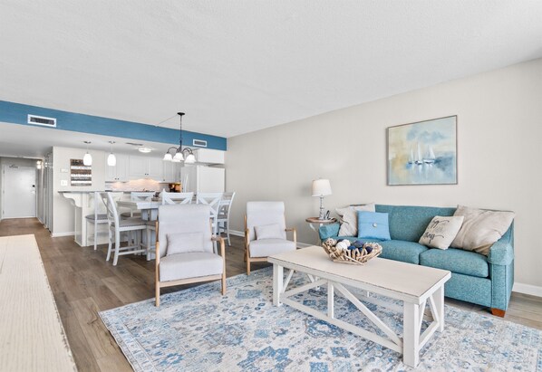 A modern living room with a blue sofa, white armchairs, a coffee table, and a dining area with white furniture. - Garden City Beach Oceanfront Condo 912 | Rising Tides Properties Rental (Murrells Inlet)
