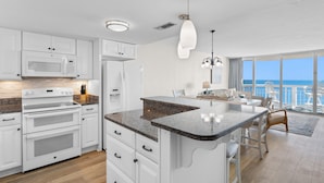 A modern kitchen with white cabinetry, a granite countertop, and a central island. There is a dining area with a view of the ocean. - Garden City Beach Oceanfront Condo 912 | Rising Tides Properties Rental (Murrells Inlet)