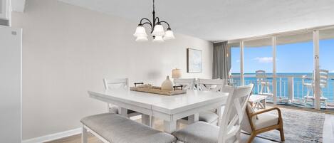A dining area with a white table, grey cushioned chairs, a chandelier, and a view of the sea through large windows.