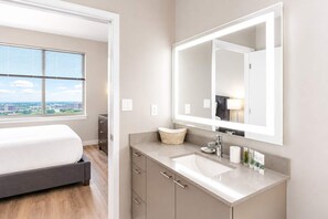 A modern hotel room with a bed, a large mirror, a vanity with a sink, and a view of the city through the window. - Luxury Apartment In Harbor Point (Stamford)