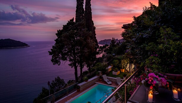 Vista della piscina con un tramonto sul mare, immersa nel verde e tra cipressi.